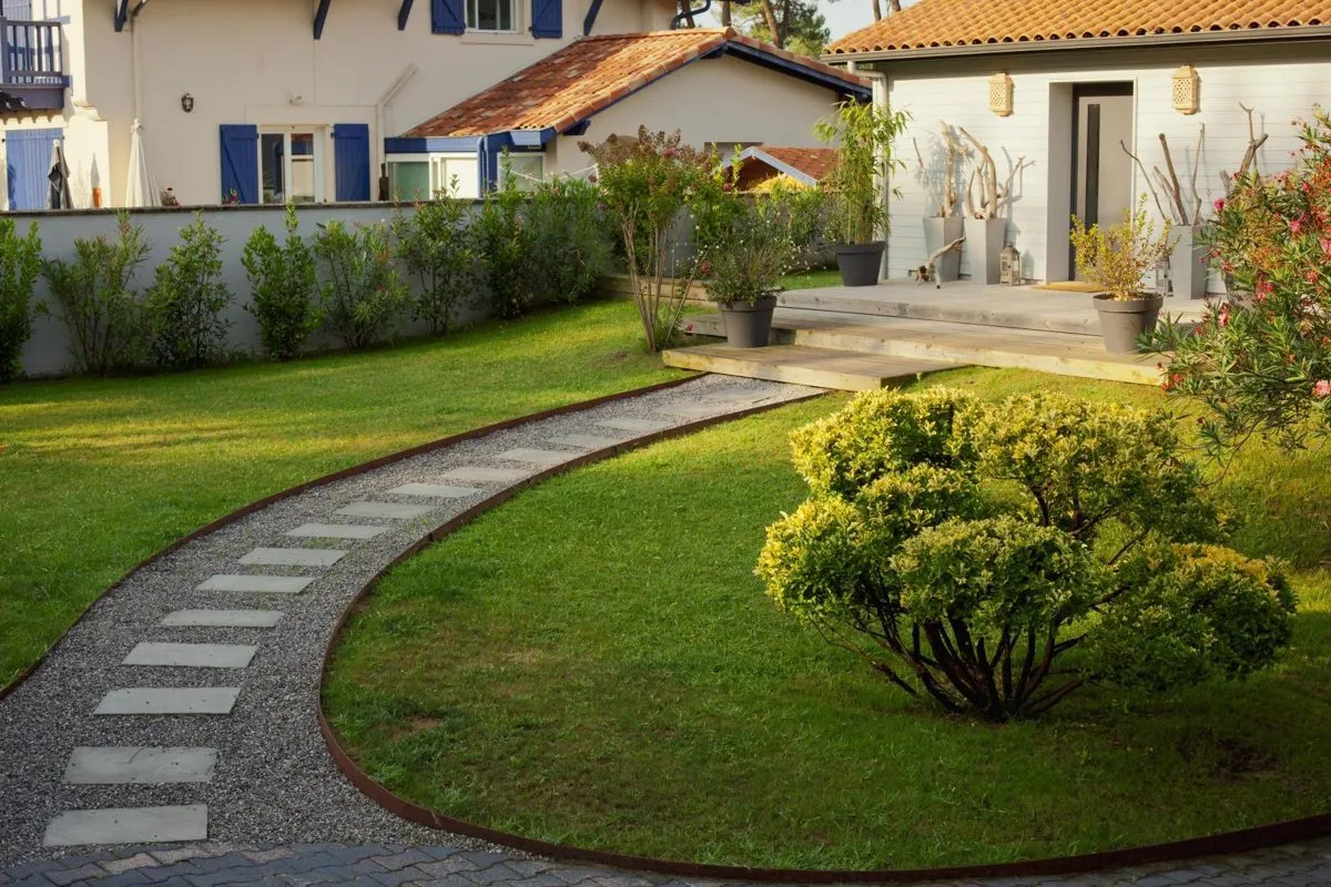 Jardin paysager au Pays Basque : allée en pas japonais, massifs et haie taillée devant une maison à volets bleus